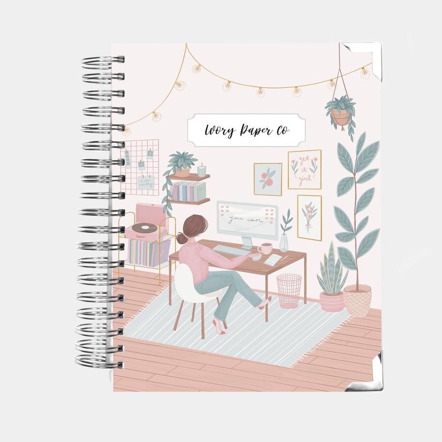 The cover of a spiral bound planner from Ivory Paper Co. The image on the cover features a woman sitting at a desk working at her computer. She is surrounded by plants, posters, and books in a soft pink, green, blue, and brown tones.